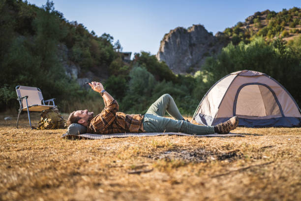 Adult caucasian man lying down near tent and use mobile phone on camping trip